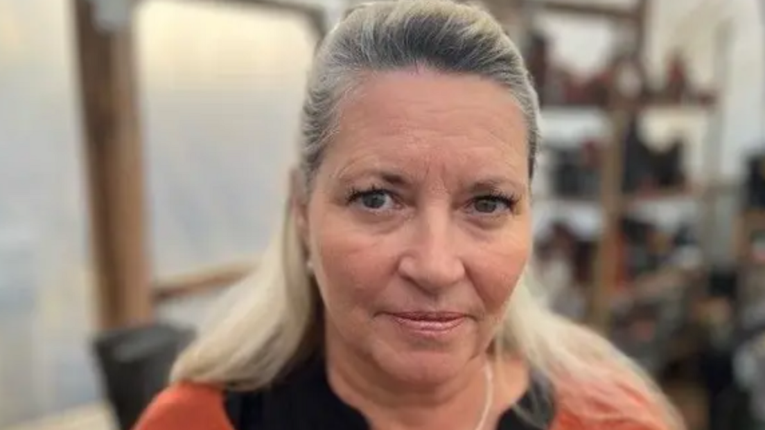 A woman with long blonde hair looks at the camera. She is earing an orange and black top and a gold necklace. Behind her, blurred, is a window and shelves