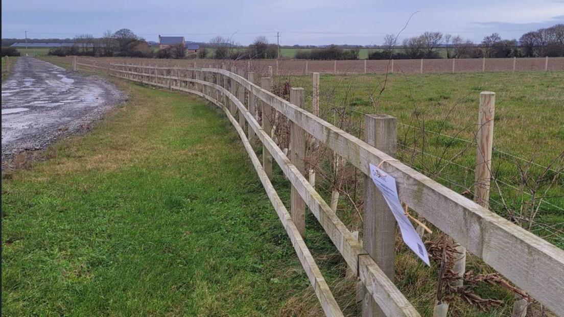 A wooden fence cuts across fields into the distance. A piece of paper has been attached to the fence, but the writing cannot be seen.