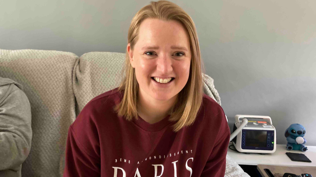 A woman with shoulder length blonde hair smiles at the camera. She is wearing a burgundy top with the word Paris on it