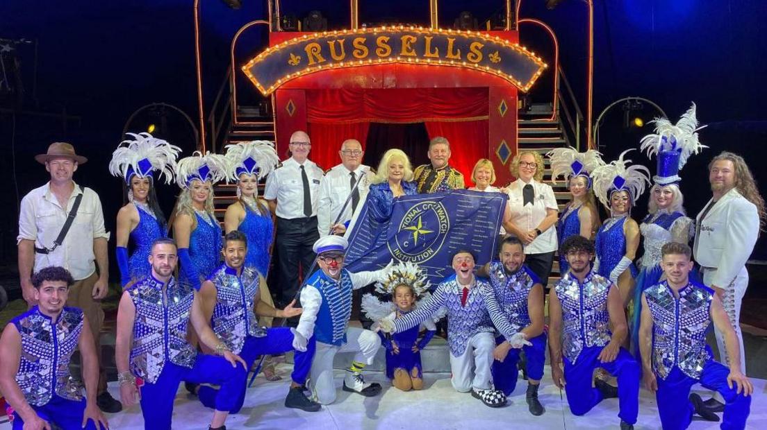 Circus performers dressed in blue and white outfits with ruffled white feather hats on the women holding the flag