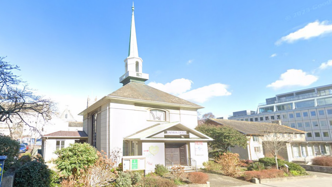 Plymouth Unitarian Church is a square white building with large wooden doors and with a spire that has a balcony. It is detached and sits back from a road. It has mature shrubs and small trees planted around the entrance path. There are tall office blocks nearby. It is a bright blue sky day.