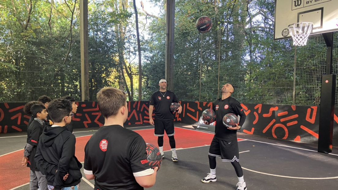 Several boys and two men wearing all black stand on an outdoor basketball pitch. One man holds two black and red basketballs while another is thrown in the air above him.