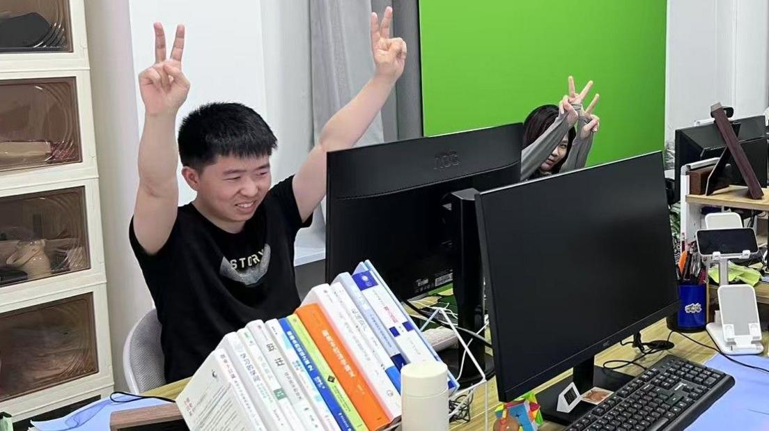 Shui Zhou, a young adult who pretends to have an office job, makes the victory sign with both hands that he is holding up in the air