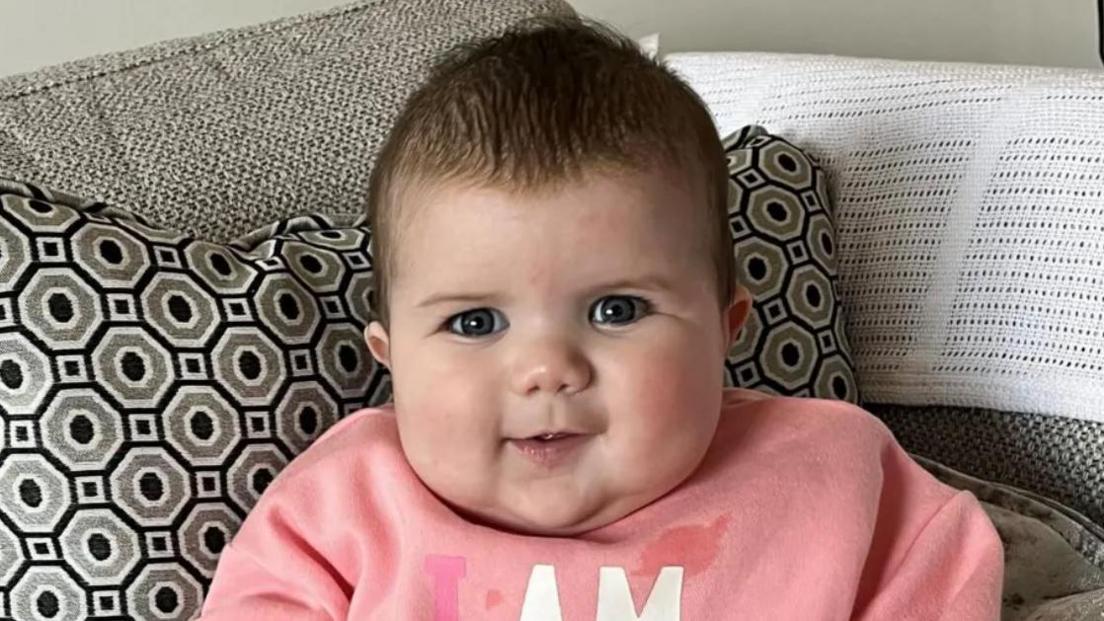 Mabli Hall sitting propped up in a grey sofa with black and grey patterned cushion. She is wearing a pink jumper