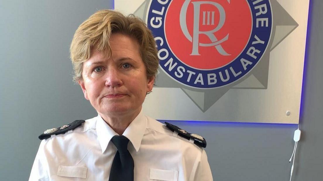 Maggie Blyth, who has short light brown hair, looking straight ahead. She is wearing her uniform of a white shirt and black tie with black patches on her shoulders. The logo for Gloucestershire Constabulary is behind her on the wall.