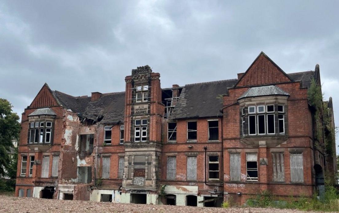 A derelict former hospital building in red brick with lighter coloured stone detail including first-floor bay windows and a plaque reading Eye Infirmary. Ground flood windows have been boarded up, and parts of some walls demolished. The roof has gaping holes in the tiles.
