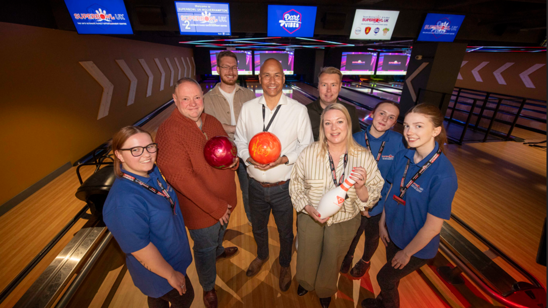 A group of people are standing on a lane in a bowling alley. Two of the men are holding bowling balls and a woman is holding a skittle.