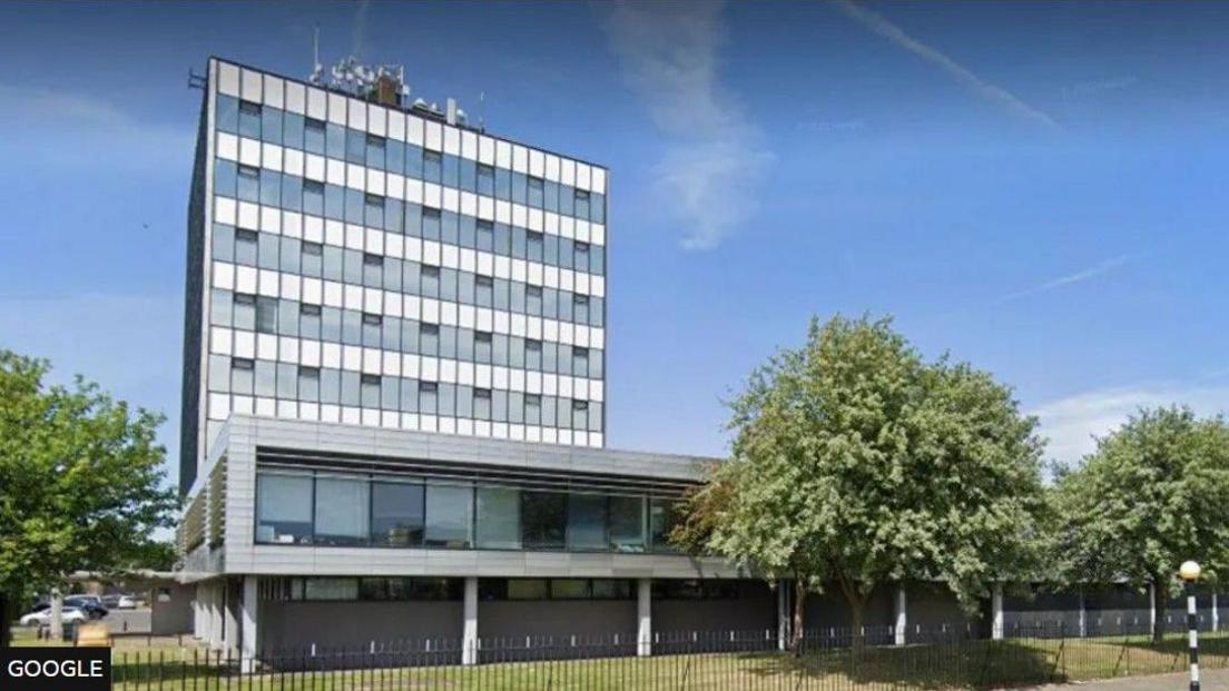 A street view of Halton council's offices. The offices are contained in a tower block which is surrounded by trees and railings, with a car park to the left