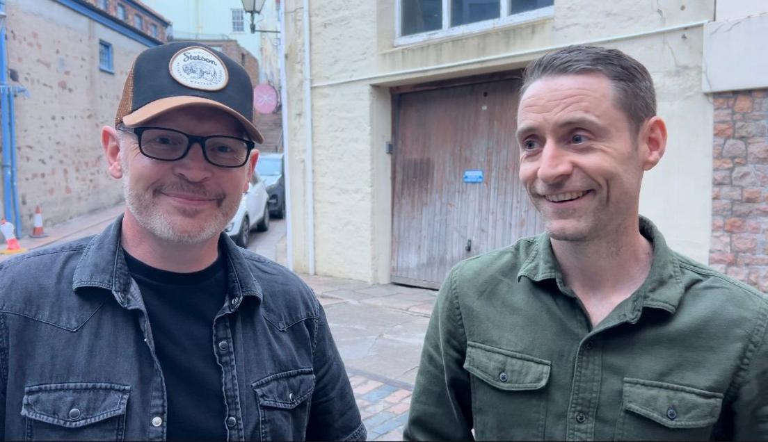 Steve Doyle is wearing a cap and glasses and a black denim jacket. Barry James stands on his right wearing a green shirt.