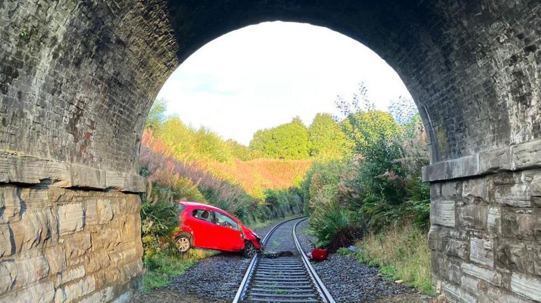 A red car which has gone down an embankment and is lying with the front wheel beside a railway track