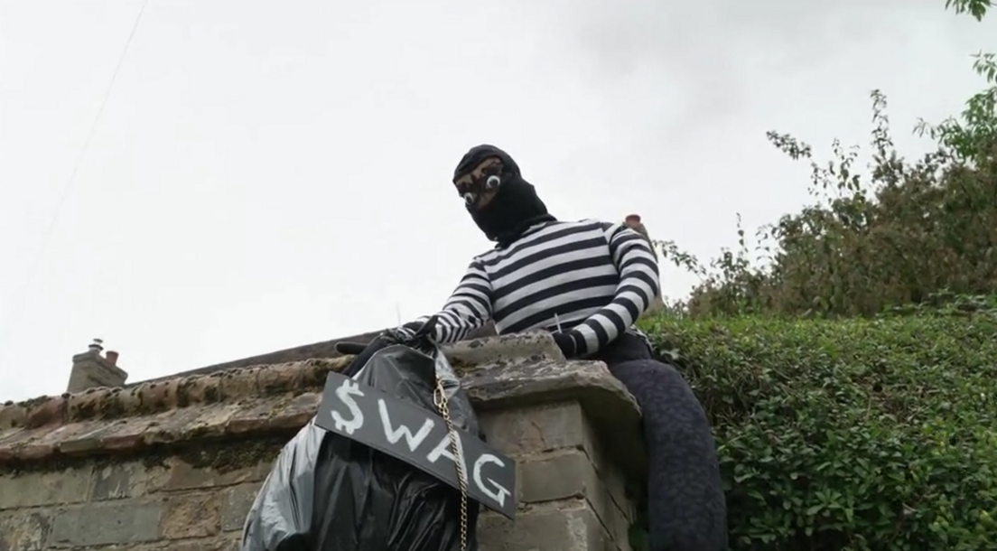 A burglar scarecrow is climbing over a wall and holding a black binbag with a sign that says 'swag'. It is wearing a black and white striped top and balaclava. 