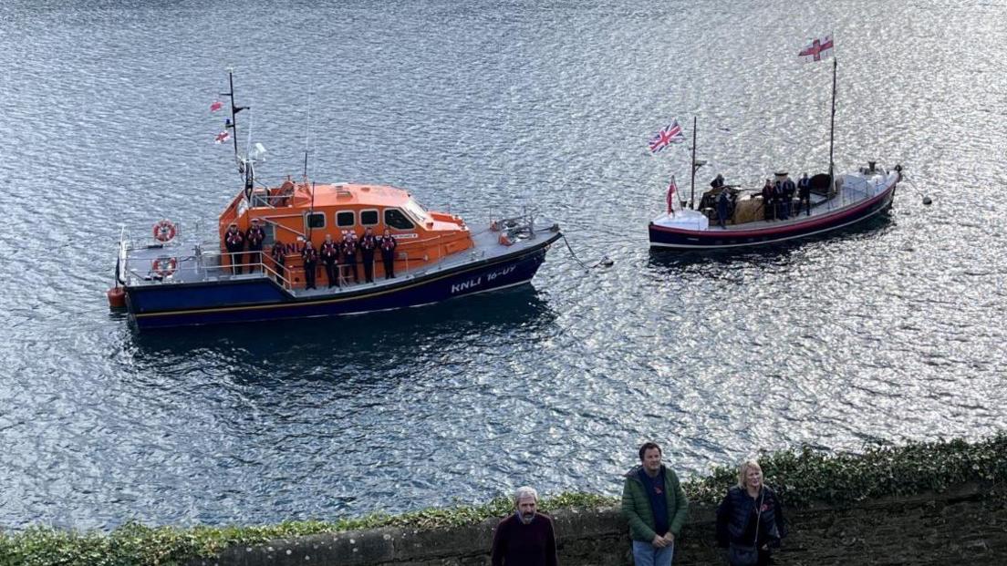 Two boats including one from the RNLI on the water in Salcombe with their crew holding poppy wreaths during a Remembrance Sunday service.