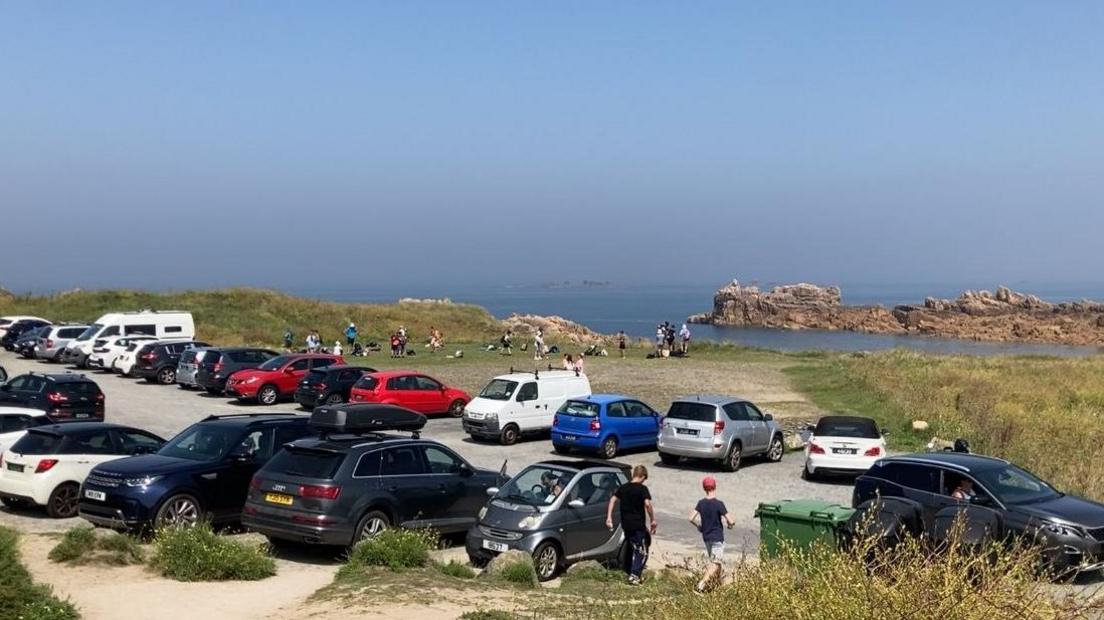 View of Grandes Rocques beach car park, looking towards the sea. There are numerous cars in the car park.