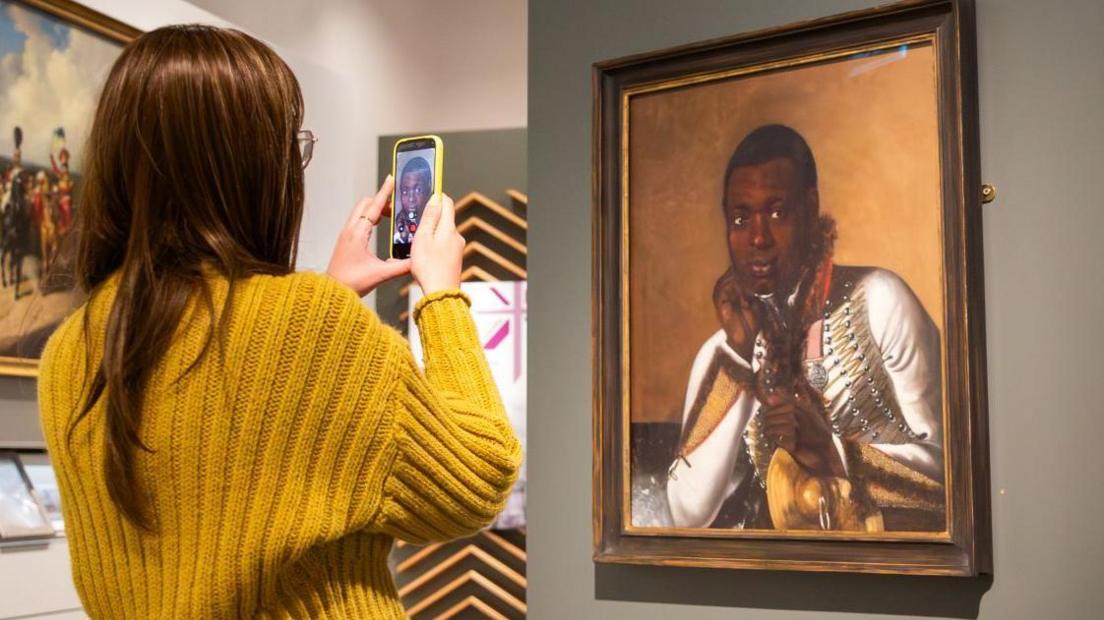 A woman wearing a yellow jumper takes a photo of a painting. The work depicts a black military bandsman holding a cymbal.