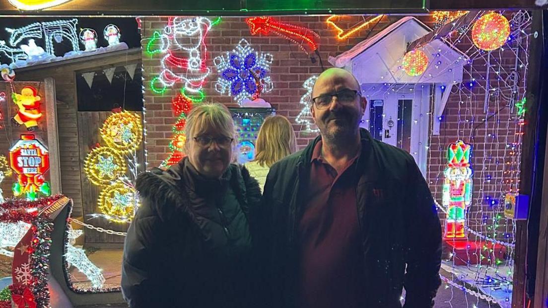 Two adults stand outside a house covered in colourful Christmas lights and decorations. Bright neon shapes, Santa figures and glowing ornaments fill the background.