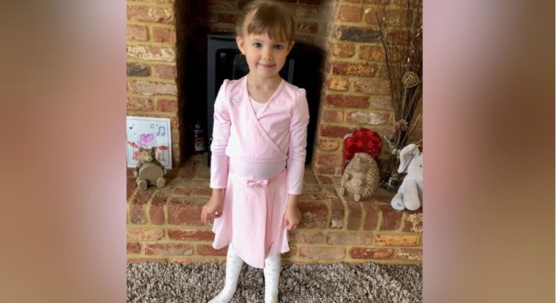 Jessica in a pink ballet outfit, with brown hair, her hair tied back, standing in a living room in front of a brick fireplace.
