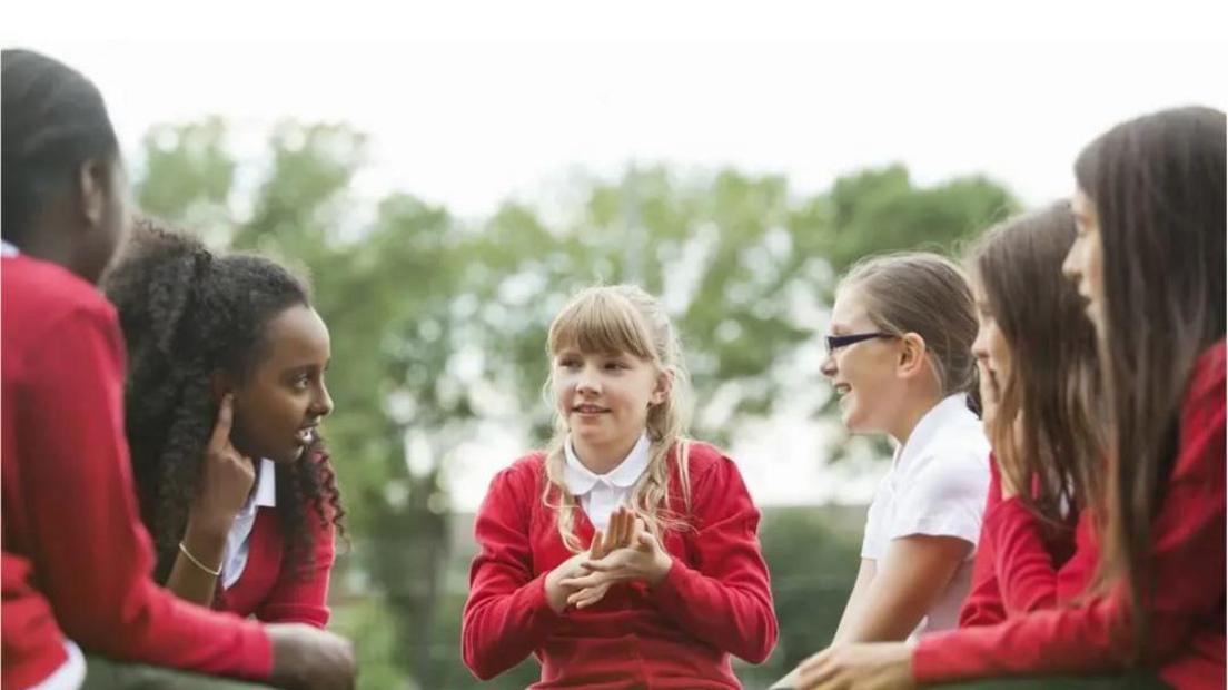 Five school children in red uniform sit around talking to each other with a backdrop of blurred trees. Stock image