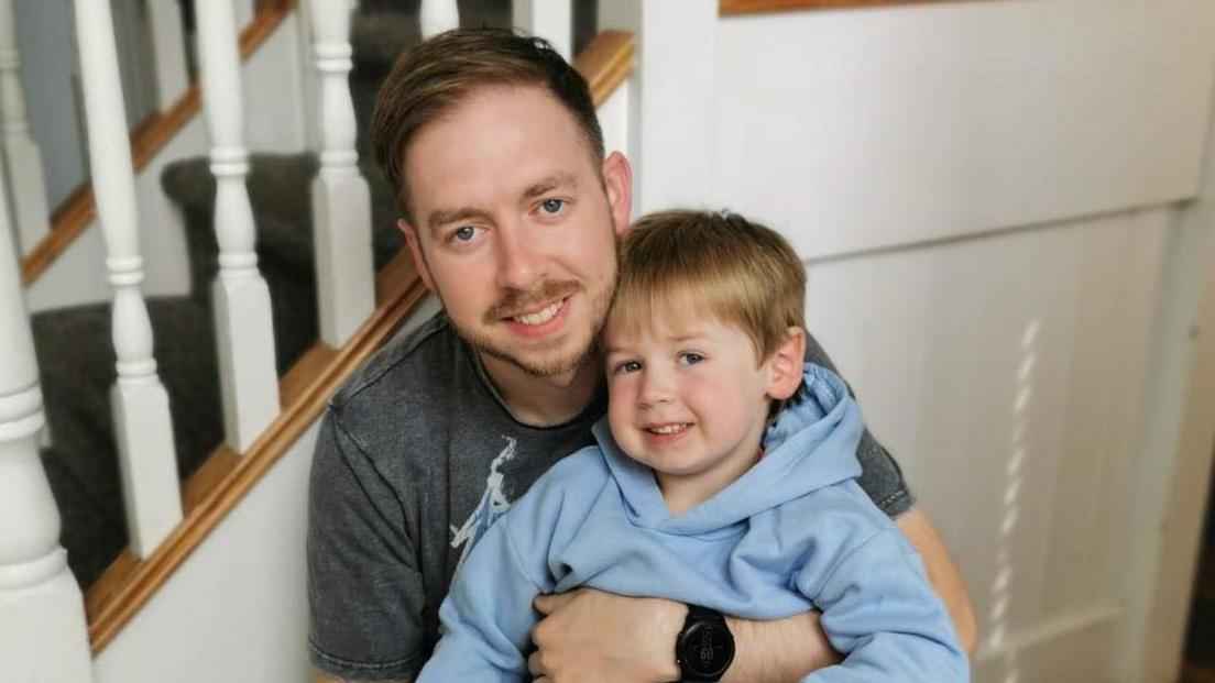 Chris has brown hair. He's a short moustache and beard. His son is wearing a blue jumper. They're leaning against banisters.