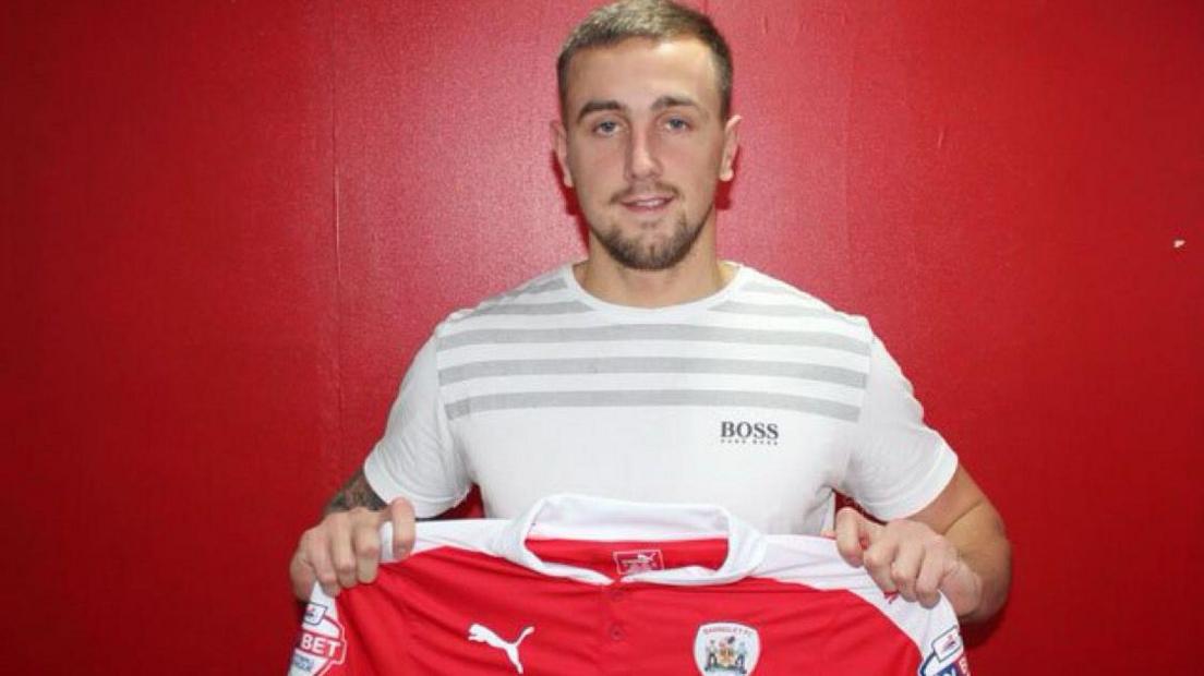 A young man in a white t-shirt holds up a red football shirt