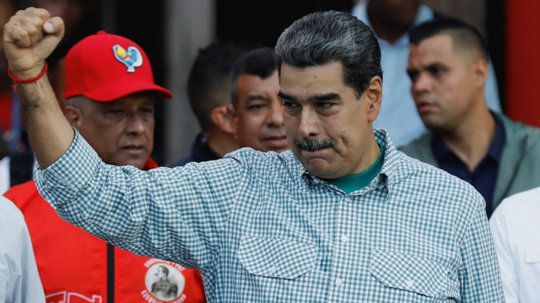 Nicolás Maduro wearing a checkered shirt hold up his right fist. Behind him, the interior minister, Diosdado Cabello, can be seen wearing a red jacket and a red baseball cap. 