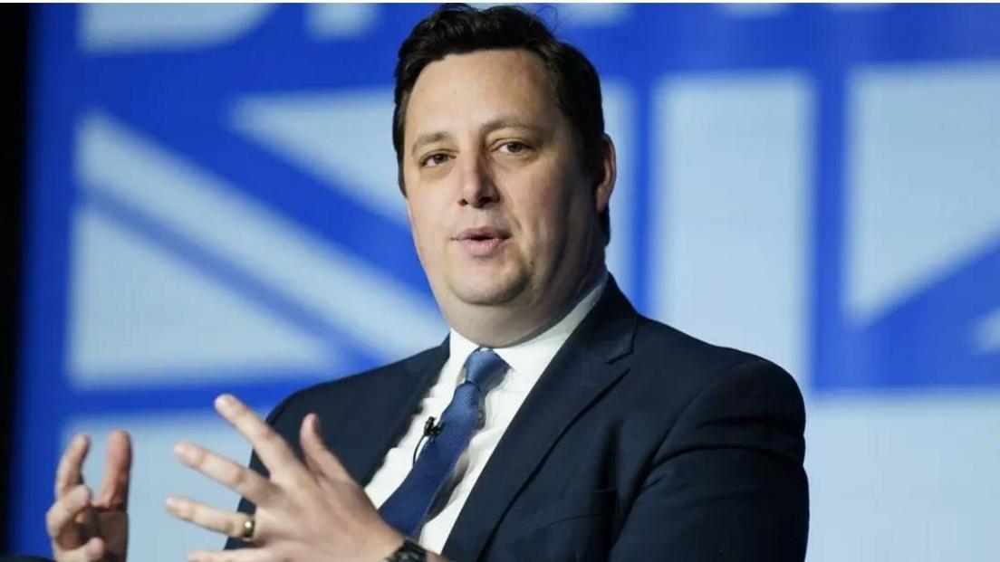 Houchen standing in front of a blue and white background with his hands held up. He has dark hair and is wearing a jacket and blue tie.