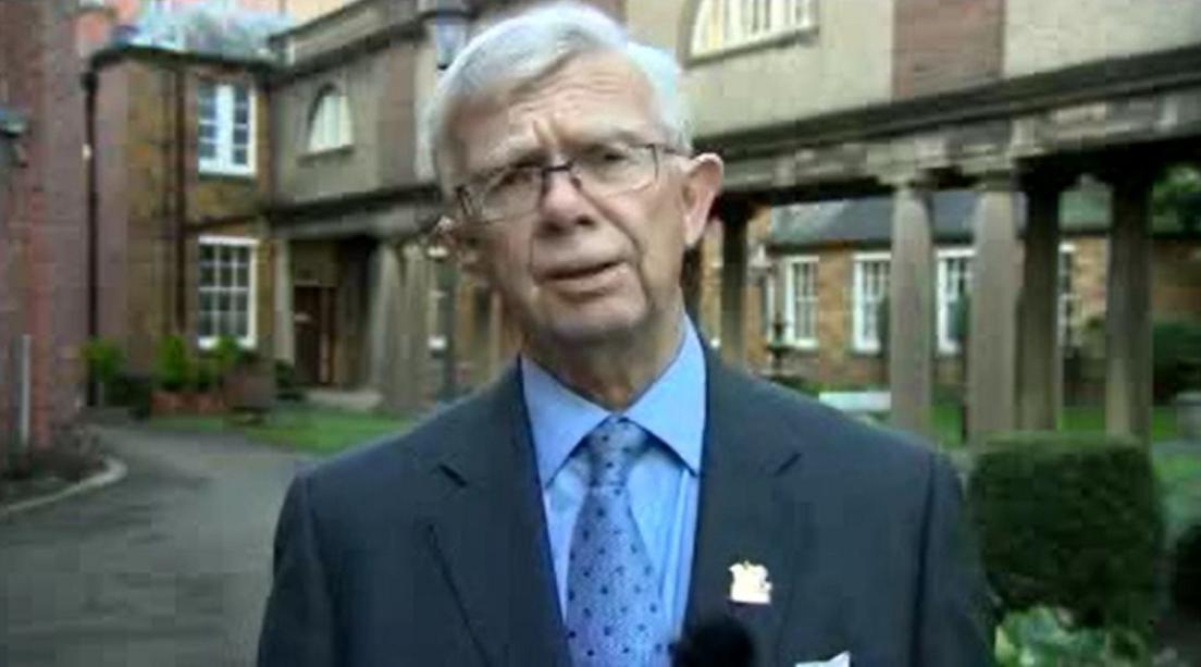 Jim Harker with white hair and a moustache wearing a grey suit and blue shirt and tie outside a two-storey office building.