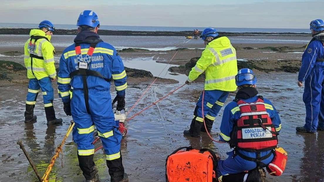 Several workers in high-vis jackets on a muddy beach with winching materials helping rescue a walker