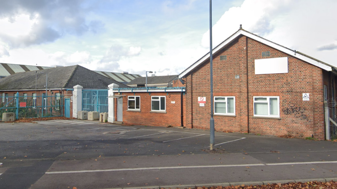 A brick building. There are four parking spaces and a lamppost outside and blue metal fencing around parts of the building.