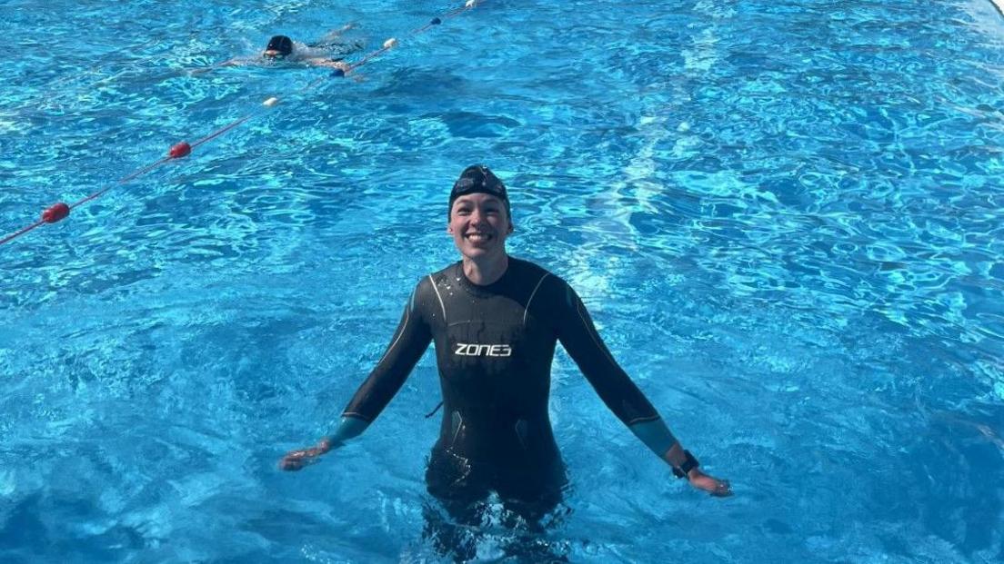Lucy is standing in the lido withe the water up to her waist and her arms stretched out to her sides. She is smiling at the camera and the water is a bright blue. She is wearing a black wetsuit - with Zones written in the centre - and a black swim cap with goggles on her head.