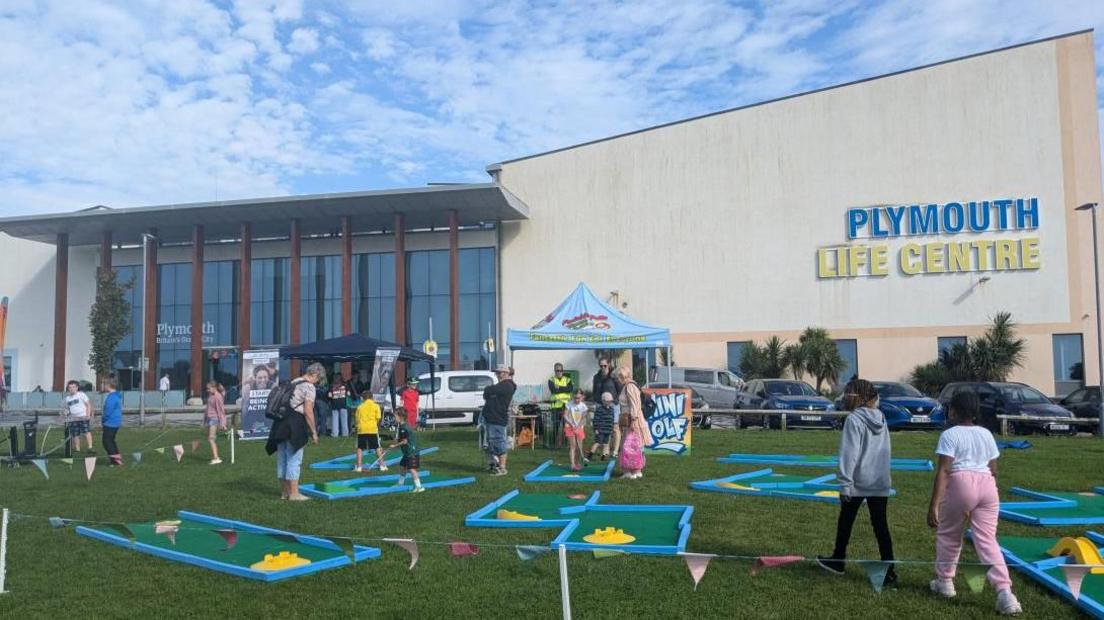 A mini golf course on the grass outside the Plymouth Life Centre in Plymouth Central Park. There are a number of different courses with lots of children take part. There is the big Life Centre building in the background with clear blue skies. 