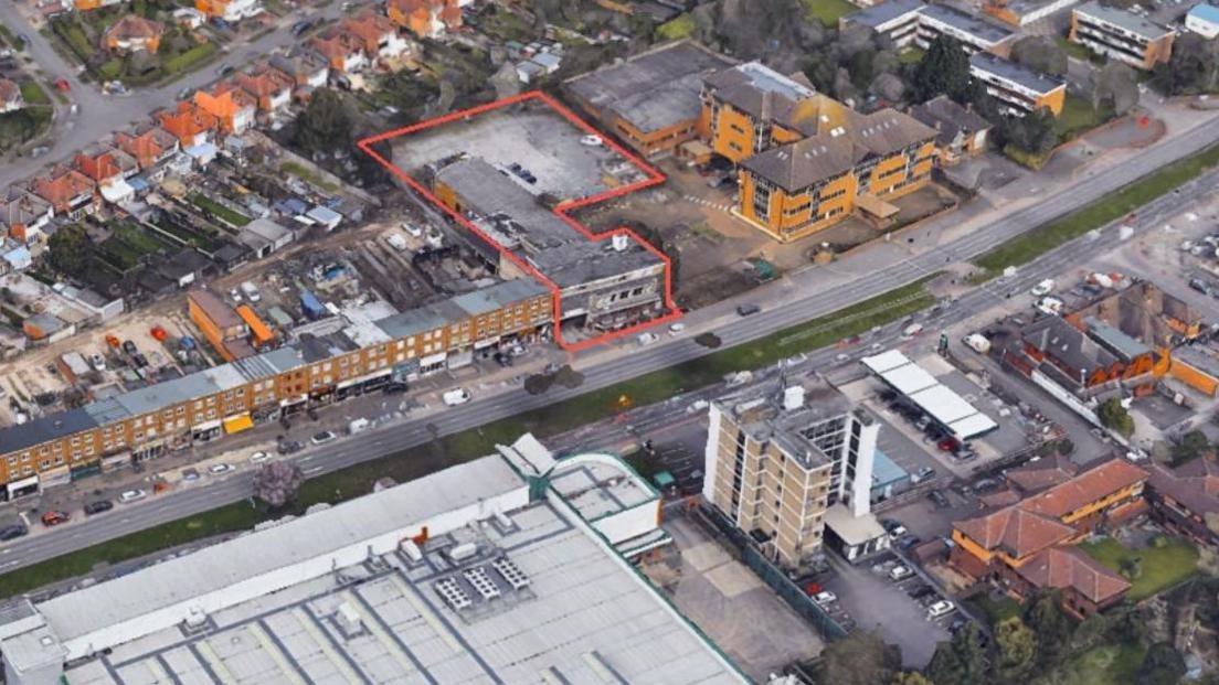 An aerial image of the site, which is marked out by an orange line. Other buildings are to the left and right of the site on the road.