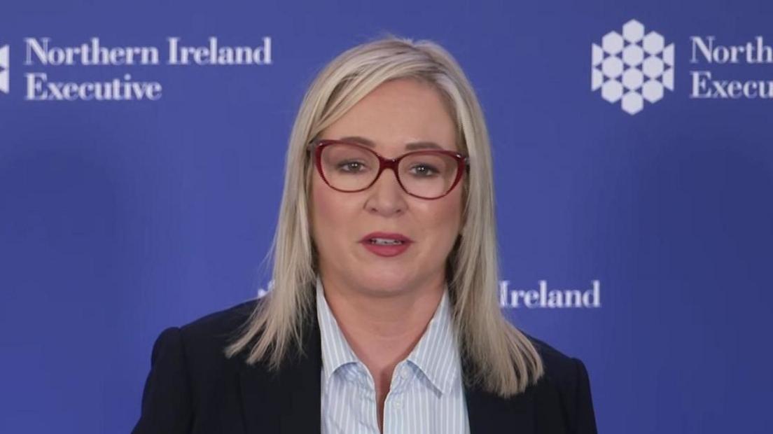 Michelle ONeill in a blue blazer and striped blue top. She stands at a mic. She has blonde hair and glasses.