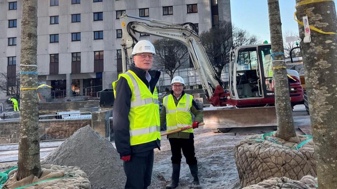 Two men wearing hard hats and hi-vis clothing in front of a construction site. In the background is a digger and next to them are trees with their roots in brown sacks.