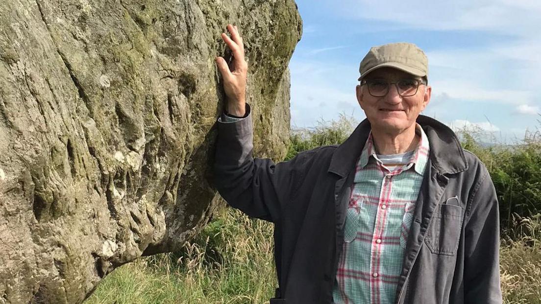 Christopher Neilan is wearing a checked green and red shirt and thin black jacket over it. He is wearing a light brown baseball cap and glasses and has one hand on a massive rock outcrop right next to him. He's somewhere outdoors with wild scrubby grass behind him. The sky is pale blue with wisps of clouds. 