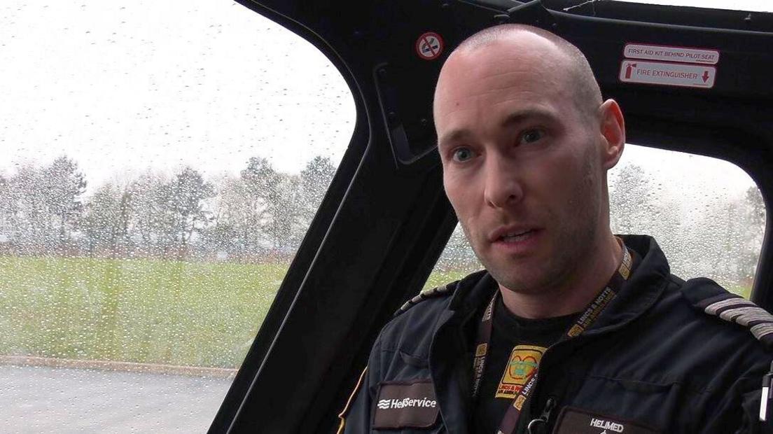 A man in black pilot overalls sits inside a helicopter cockpit with rain streaks on the windows.