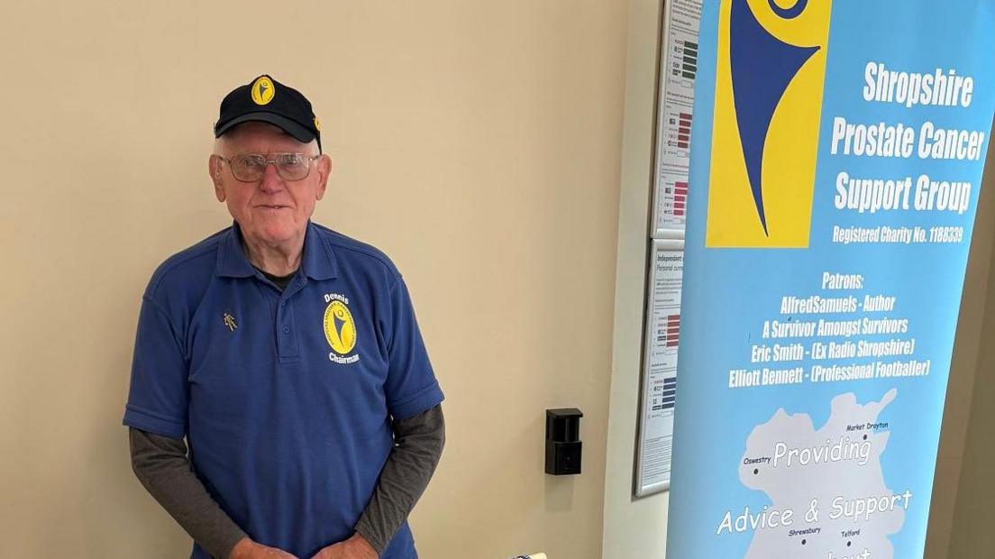 An older man with glasses is wearing a black hat with a yellow logo on it. He has glasses and is wearing a dark blue t-shirt with the yellow logo on it, and grey long sleeves underneath. Next to him is a sign that reads "Shropshire prostate cancer support group"