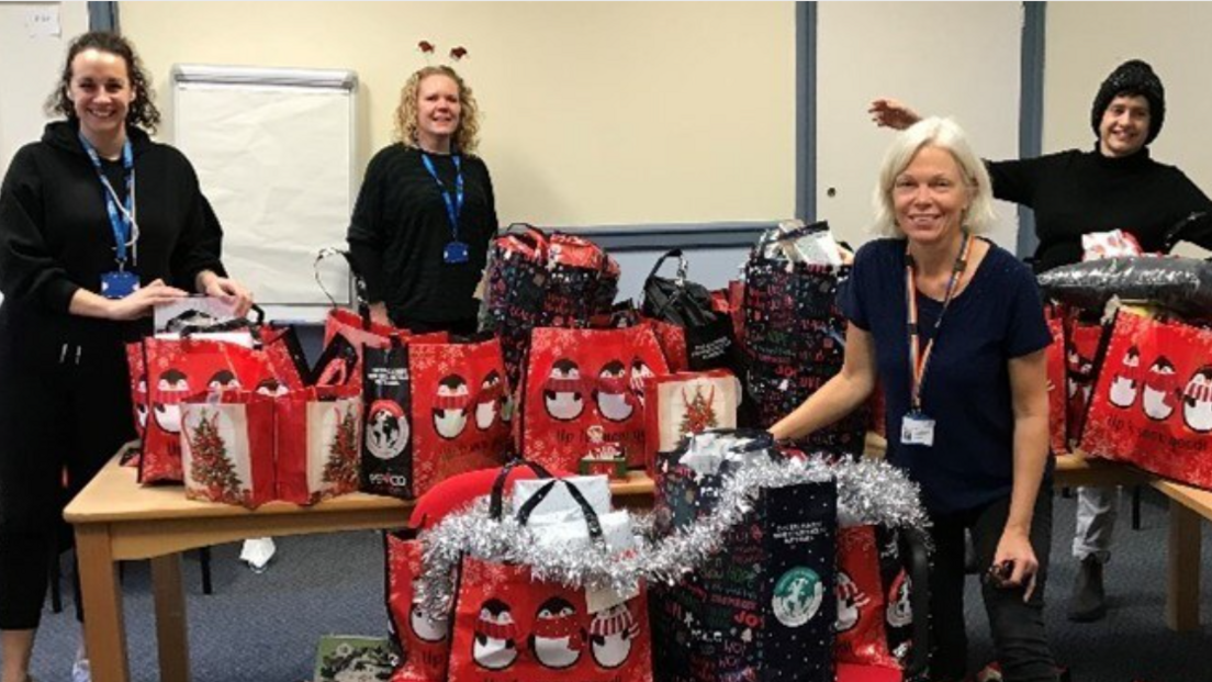 Four New Leaf workers stand with shopping bags decorated with pictures of Christmas trees and penguins under a silver tinsel garland