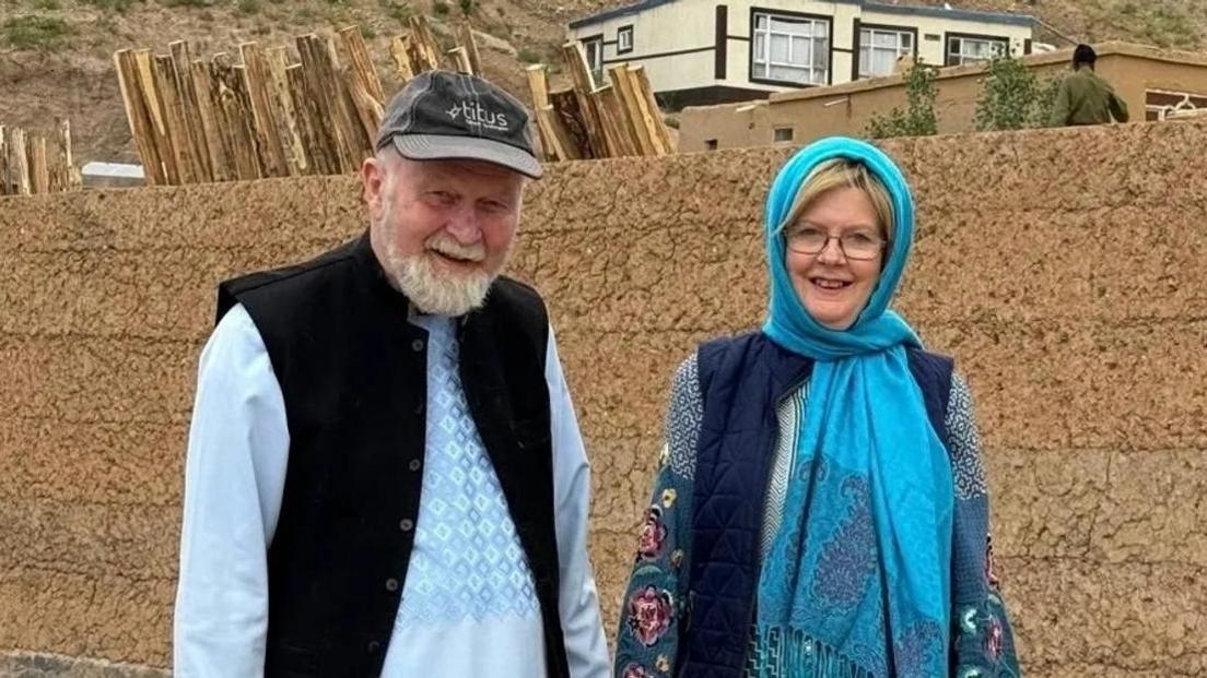 Peter and Barbie Reynolds smile at the camera in front of an orange brick wall. Peter is an elderly man wearing a blue long-sleeved top, a black waistcoat and a grey cap, while Barbie wears a bright blue headscarf, a navy gilet and a patterned blue top.