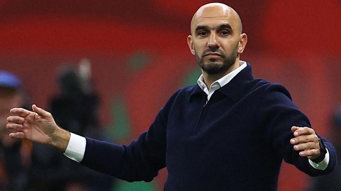 Walid Regragui, wearing a blue zip-neck jumper over a white shirt, spreads his arms out while showing a neutral expression during a football game