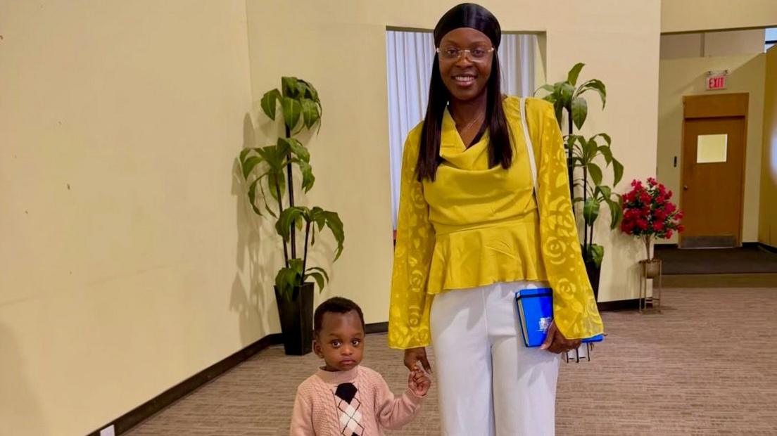 Ngozi Okobi-Okeoghene wears a mustard-yellow blouse, white trousers, a black headwrap and glasses while holding a blue book at her left hip. With her right hand she is holding hands with little boy dressed in a light pink sweater. Both are looking into the camera lens. The setting is a spacious, carpeted indoor lobby featuring neutral-coloured walls, two potted green plants, a red floral arrangement and what looks like a fire door in the background.