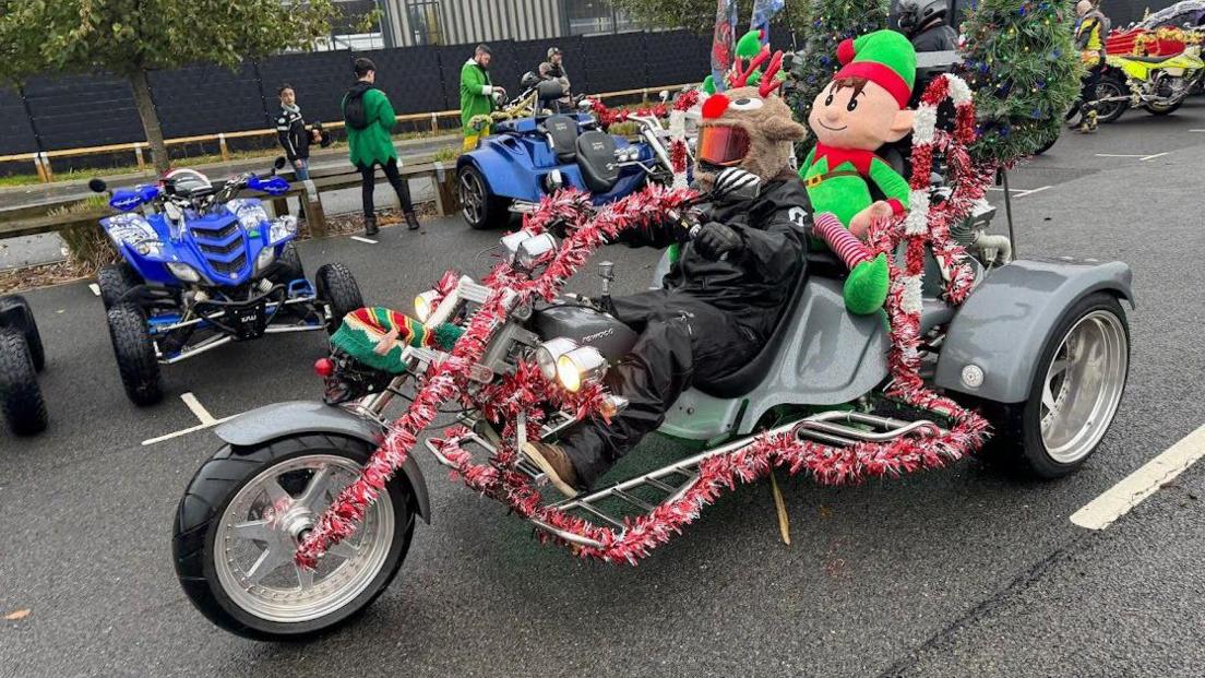 A grey trike covered in tinsel. The rider has a reindeer head on his helmet. His pillion passenger is a giant elf soft toy.