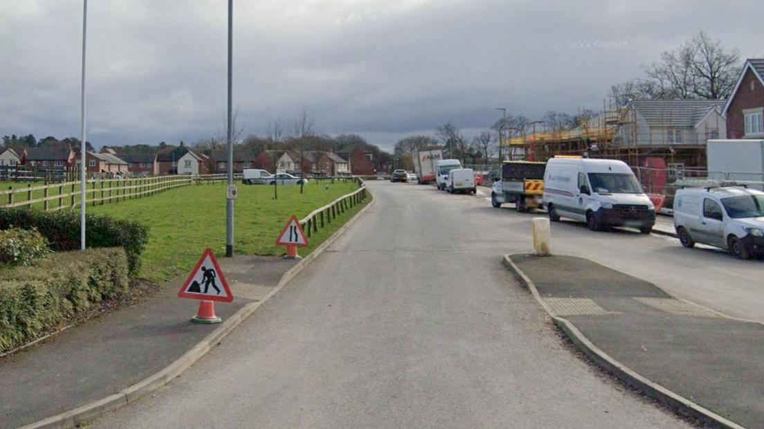 A plot of land off Henry Littler Way in Whittingham. A green area can be seen to the right of the road, with parked vehicles on the left. Housing can be seen in the background.