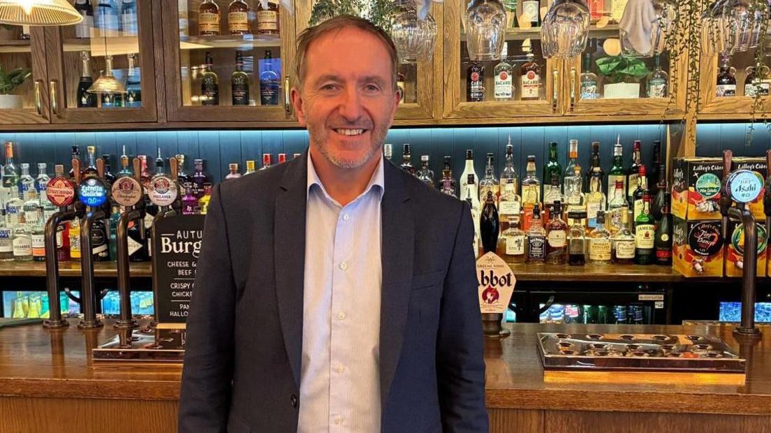 Pub boss Nick Mackenzie, dressed in a casual suit, stands in front of one of his Greene King pubs. The wood-panelled bar has a selection of beers on draught, a fully stocked bar in the background also has some Halloween decorations