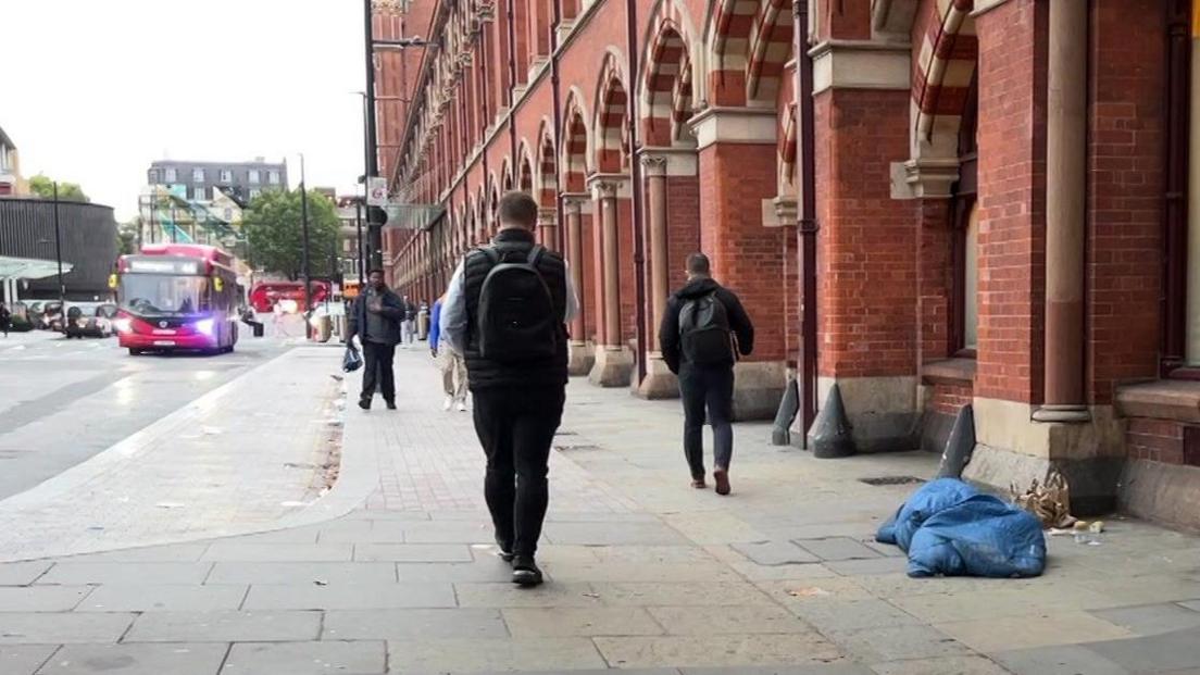 A rough sleeper in a blue sleeping bag near Kings Cross station as people walk by