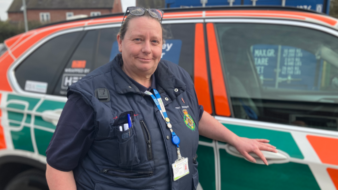 A woman in a blue T-shirt and blue gilet stands alongside an emergency vehicle, smiling. She has glasses on the top of her head, and an NHS lanyard around her neck and some pens in her picket.