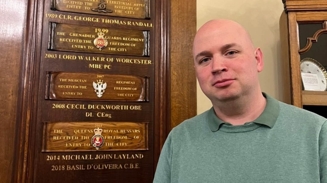 A bald man in a green jumper standing in front of a number of wooden plaques mounted on a wall