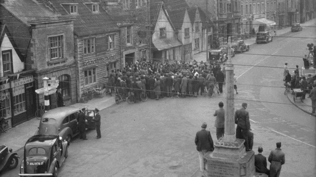 A black and white photograph taken from above looking across a square over a war memorial toward a large crowd of well dressed people pressed up against the entrance of a hotel, as a film crew opposite watch events unfold and smart classic cars line the street.