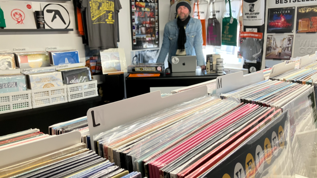 A man is stood behind a counter leaning over a silver laptop with an apple logo on the front. He is wearing a denim jacket and dark scarf tied around his neck, and in the foreground is a range of vinyl in racks, ordered alphabetically.