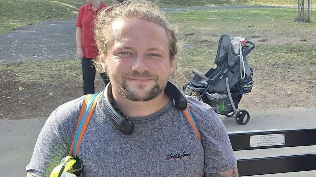 Ashley Southall sat on a bench ready for his walk. He has a sports top on and a fan around his neck. There is a pushchair behind him in the background. He is smiling and has long hair and a goatee-style beard.