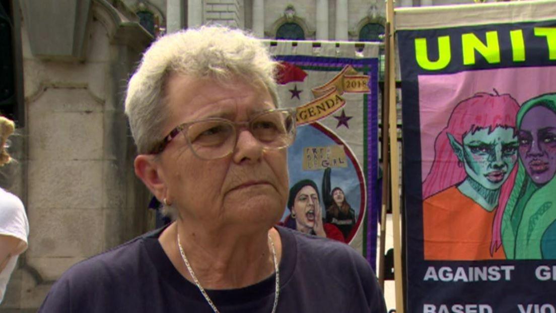 A woman with short grey hair, pictured from the shoulders up. She is wearing a navy top, a necklace and glasses. There are anti-racism posters hung up behind her.